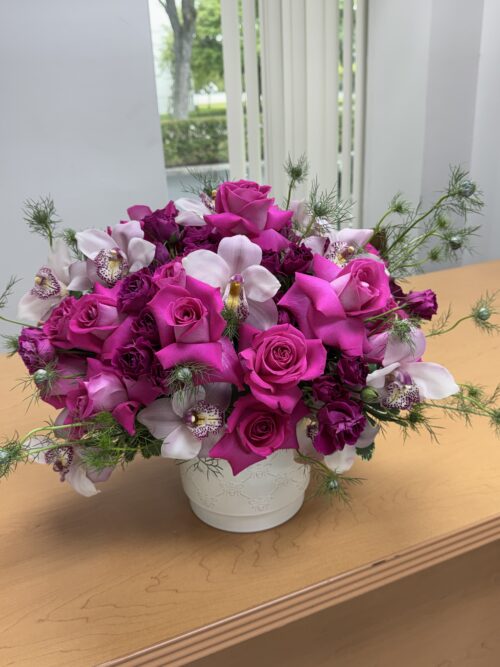 Bright pink rose and orchid bouquet in a white vase on a wooden table, with light background.