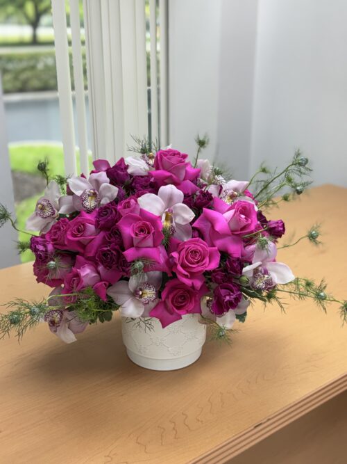 Bouquet of bright pink roses and pink orchids in a white vase on a wooden table by a window with blinds behind it