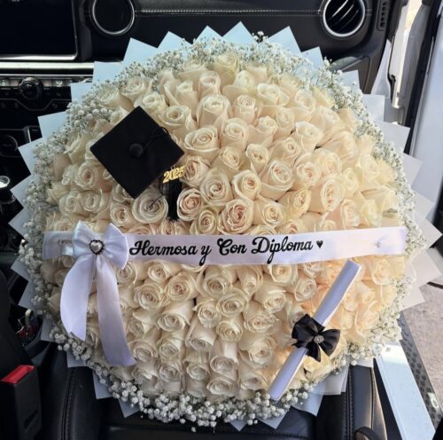 Circular bouquet of cream roses with baby's breath, a black graduation cap, and a white ribbon reading 'Hermosa y Con Diploma' beside a rolled diploma in a car interior