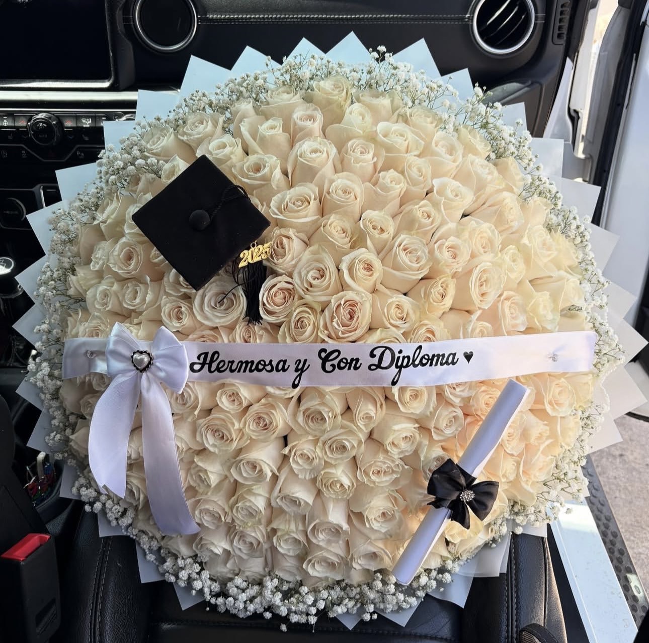 Circular bouquet of cream roses with baby's breath, a black graduation cap, and a white ribbon reading 'Hermosa y Con Diploma' beside a rolled diploma in a car interior